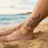 Woman's tattooed ankle resting on golden beach sand with ocean blurred in background