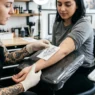 tattoo artist holding printed reference design against woman's wrist checking size in bright studio
