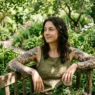 Woman with botanical tattoos in lush garden setting with dappled sunlight on tattooed arms