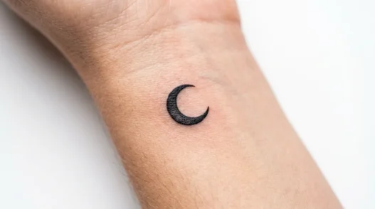 Tiny crescent moon tattoo on woman's inner wrist with clean black line work and precise details