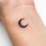 Tiny crescent moon tattoo on woman's inner wrist with clean black line work and precise details