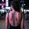 Woman's back showing intricate cybersigilism spine tattoo with circuit board patterns along vertebrae in neon lighting