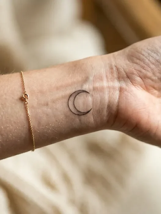 Woman's wrist showing single delicate fine-line moon phase tattoo with soft studio lighting