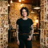 Tattooed woman standing against wall of traditional tattoo flash art sheets in authentic studio setting