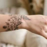 Delicate fine-line floral tattoo wrapping around woman's wrist showing botanical elements in precise black ink
