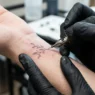 Extreme close-up of single needle creating ultra-fine tattoo lines on woman's wrist with bright studio lighting