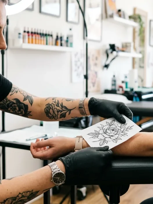 Tattoo artist holding printed flower design against woman's wrist to check size proportion in bright studio