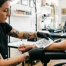 Tattoo artist holding printed flower design against woman's wrist to check size proportion in bright studio