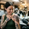 Woman looking down at fresh tattoo with delighted expression while tattoo artist adjusts protective wrap