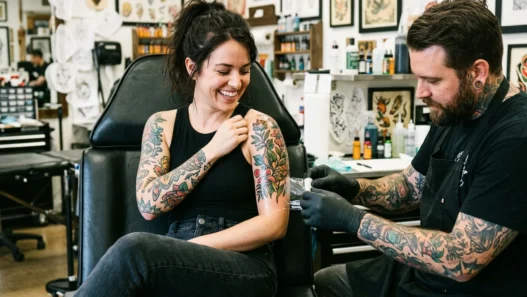 Woman looking down at fresh tattoo on shoulder with genuine delight while tattoo artist adjusts wrap nearby