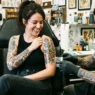 Woman looking down at fresh tattoo on shoulder with genuine delight while tattoo artist adjusts wrap nearby