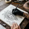 Tattoo artist's hand sketching custom Medusa design on paper with client's tattooed arm resting nearby on studio desk