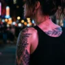 Woman with tattooed shoulder and collarbone under pink and blue neon lighting on city street at night