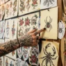 Tattooed woman's hand pointing to spider design among organized flash art sheets on tattoo studio wall
