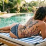 Woman on pool lounger showing spine butterfly tattoo and ribcage cherry blossoms in bikini with turquoise water background