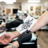Tattoo artist holding printed design reference against woman's forearm checking size proportion in bright studio