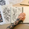 Overhead view of tattoo reference images and sketches scattered on white desk with tattooed hand resting beside planning materials
