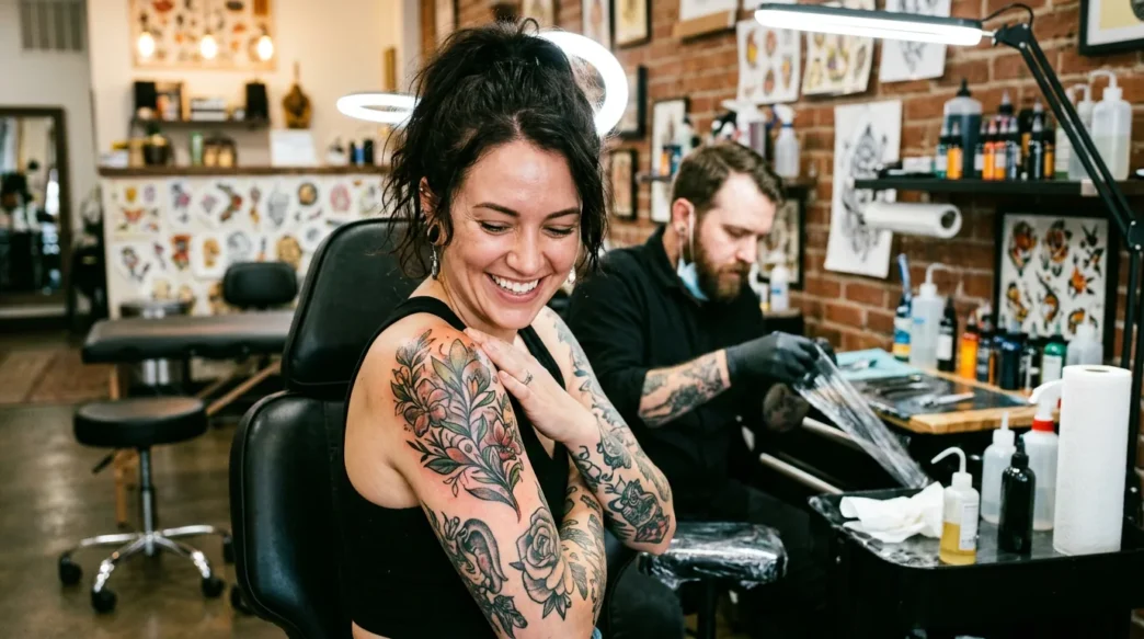 Woman looking down at fresh shoulder tattoo with delighted expression in bright tattoo studio