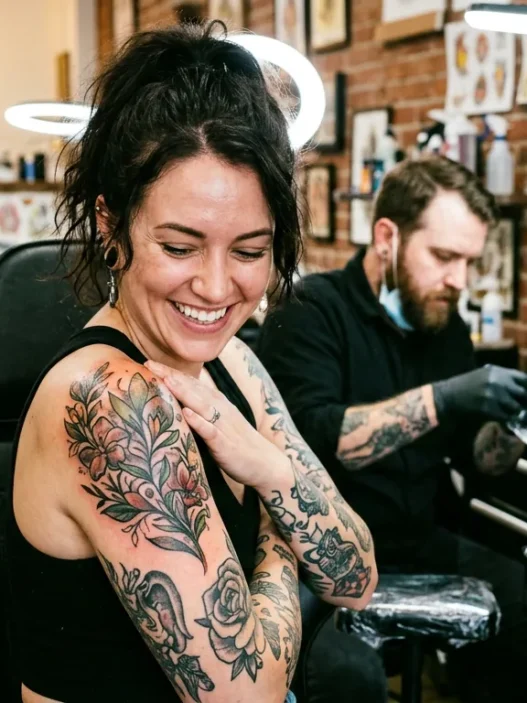 Woman looking down at fresh shoulder tattoo with delighted expression in bright tattoo studio