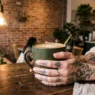Woman's tattooed hands wrapped around ceramic coffee cup in trendy cafe with warm lighting