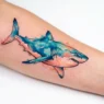 Fresh watercolor shark tattoo on woman's forearm showing blue and coral color bleeding effects