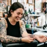 Woman in tattoo studio looking down at fresh shoulder tattoo with delighted expression while artist adjusts wrap