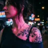 Woman with shoulder tattoo illuminated by colorful neon lights on city street at night