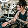 Tattooed woman in studio looking down at fresh shoulder tattoo with genuine delight while artist adjusts wrap