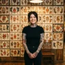 Woman with various tattoos standing in front of wall displaying traditional tattoo flash art sheets
