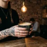 Woman with multiple finger tattoos holding ceramic coffee cup in trendy cafe setting