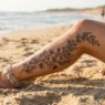 Woman's tattooed calf resting on golden sand showing a detailed botanical stem tattoo along the outer calf in coastal afternoon light