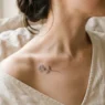 Woman's bare collarbone with a delicate fine-line rose bud tattoo against soft natural linen in warm window light