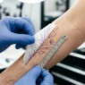 Tattoo artist positioning a fine-line stencil on a woman's outer forearm with a tape measure visible in bright studio