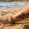 Woman's sun-kissed ankle on golden beach sand showing a bold geometric black ink shark tattoo with soft ocean blur in background