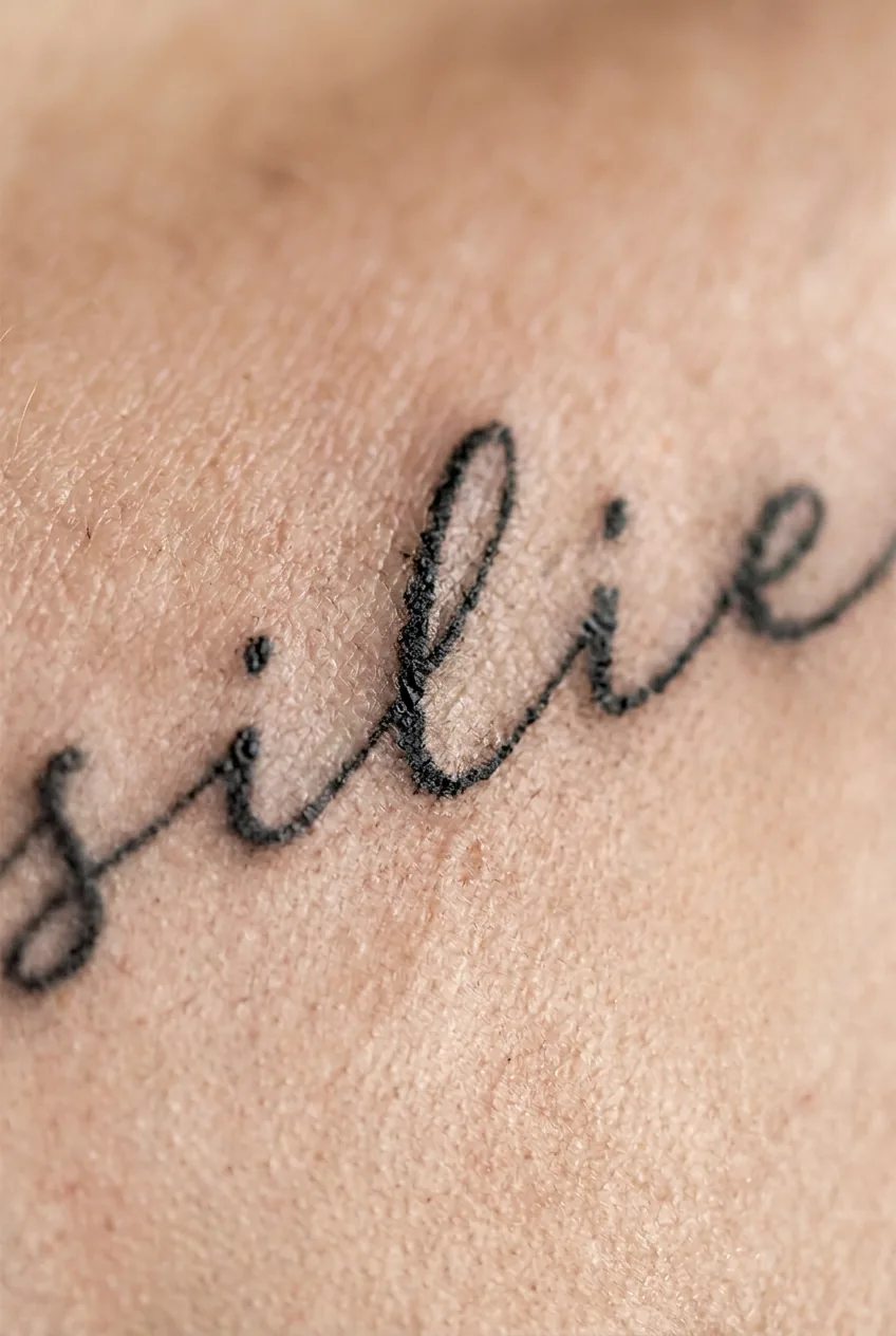 Delicate single-word script tattoo along a woman's collarbone following the bone's natural curve in soft studio light