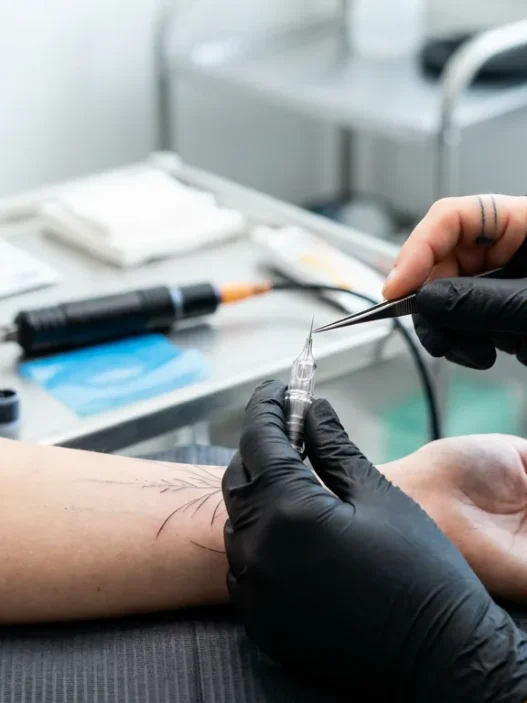 Tattoo artist preparing a single needle for fine-line work with machine resting on sterile tray in bright studio