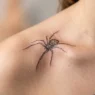 Woman's collarbone showing a delicate fine line spider tattoo with intricate leg linework under soft studio lighting