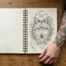 Overhead view of open tattoo sketchbook with wing designs beside woman's tattooed arm and drawing tools on wooden surface