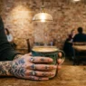 Woman's tattooed hands wrapped around ceramic coffee cup in trendy cafe with warm lighting and brick background