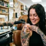 Woman with delighted expression looking down at fresh tattoo on shoulder while artist adjusts wrapping in bright studio