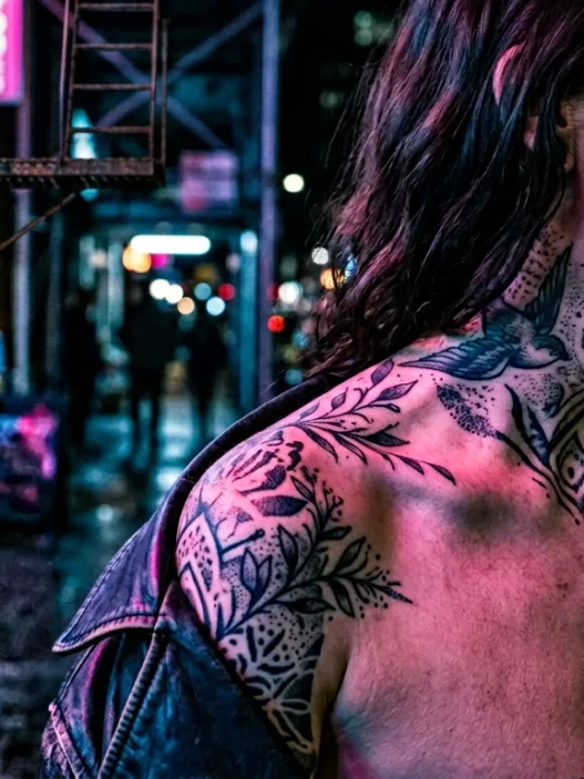 Woman with tattooed shoulder and collarbone area illuminated by pink and blue neon lighting on city street at night