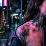 Woman with tattooed shoulder and collarbone area illuminated by pink and blue neon lighting on city street at night