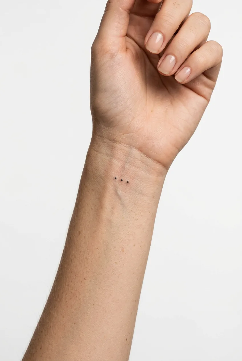 Woman's wrist featuring three tiny dots arranged as ellipsis tattoo, each dot freckle sized