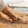Woman's tattooed ankle resting on golden beach sand with ocean blurred softly in the background in afternoon coastal light