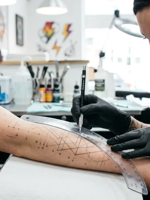 Tattoo artist using flexible ruler to plan geometric dotwork lines on a woman's forearm with pencil marks visible under bright studio light