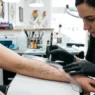 Tattoo artist using flexible ruler to plan geometric dotwork lines on a woman's forearm with pencil marks visible under bright studio light