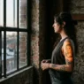 Woman with an older blurry portrait tattoo on her upper arm photographed in moody side lighting at an industrial loft window