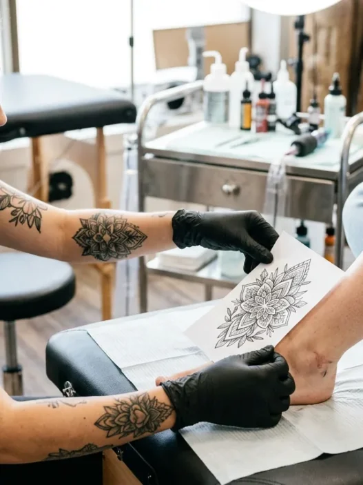 Tattoo artist holding printed reference design against a woman's foot and ankle to check size proportion in bright studio lighting