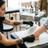 Tattoo artist holding printed reference design against a woman's foot and ankle to check size proportion in bright studio lighting