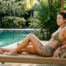 Tattooed woman in bikini relaxing on pool lounger with thigh and rib tattoos visible in afternoon sunlight
